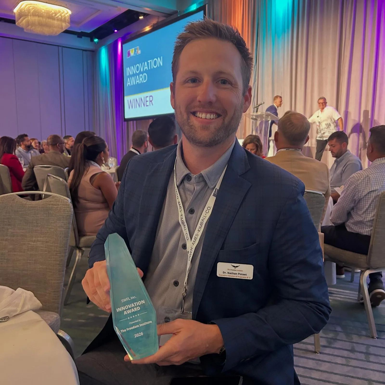 Nathan Poteet holding the Innovation Award in the ballroom, with the winner slide on screen behind him
